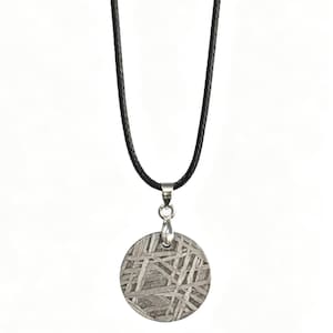 Puede incluir: Un colgante metálico circular con un patrón de superficie grabado y entrecruzado, suspendido de un cordón negro fino. El colgante está sujeto a una anilla de metal plateado, creando un diseño de collar sencillo sobre un fondo blanco liso.