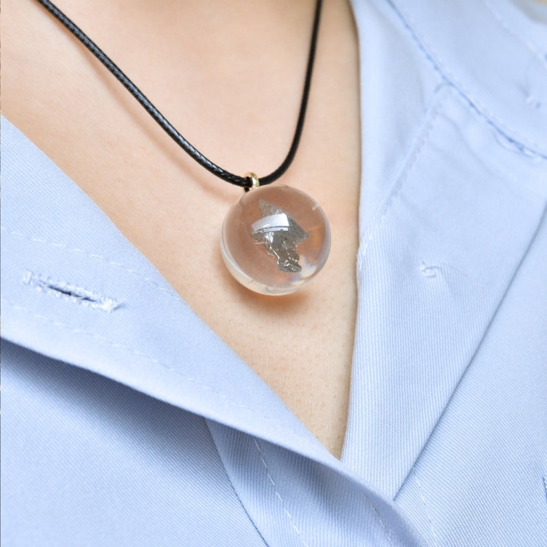 May include: A silver pendant necklace with a clear glass orb containing a small piece of metal. The necklace is on a black cord and is worn by a person with a light blue shirt.