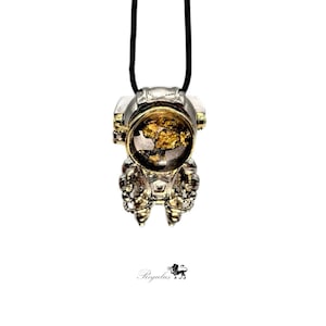 Pode incluir: Um colar com um pingente de astronauta prateado, um capacete dourado e um cordão preto. O capacete tem uma viseira transparente que revela um interior dourado.