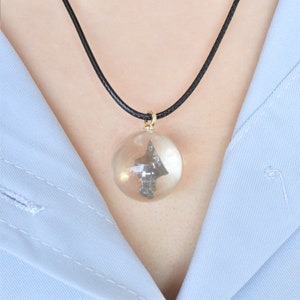 May include: A black cord necklace with a clear glass pendant containing a small, dark gray, arrowhead-shaped object.