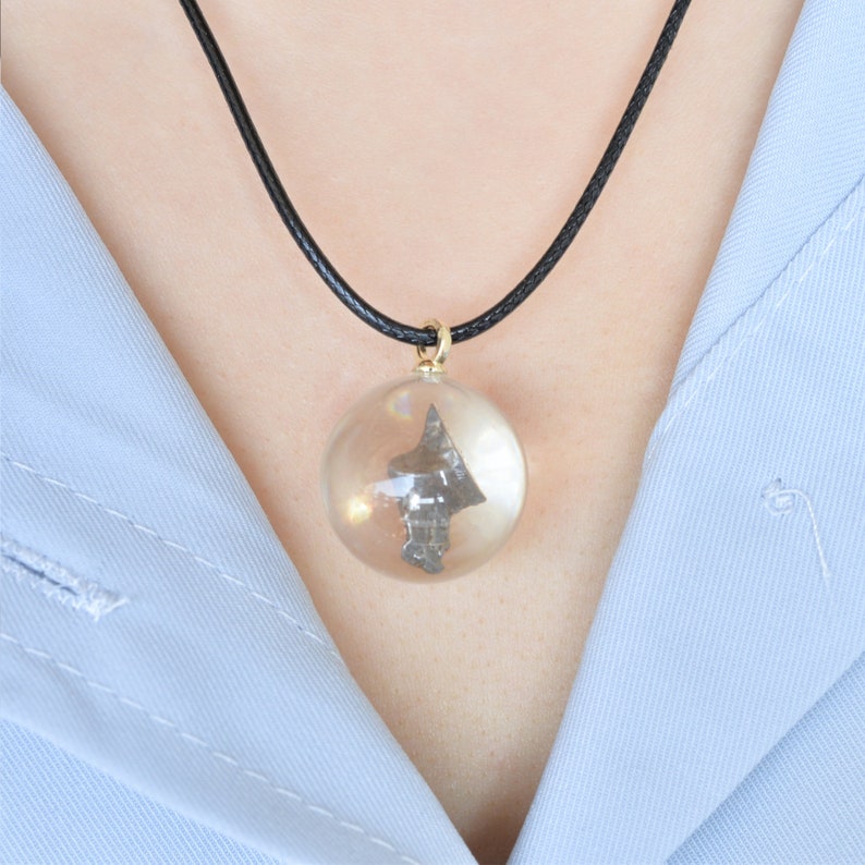 May include: A black cord necklace with a clear glass pendant containing a small, dark gray, arrowhead-shaped object.