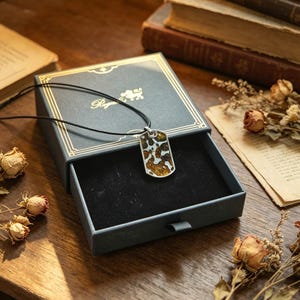 Puede incluir: Un collar con colgante rectangular con un cord&oacute;n negro, presentado en una caja azul abierta con ribete dorado. El colgante presenta un marco plateado y un dise&ntilde;o moteado en tonos marr&oacute;n y dorado. La caja est&aacute; sobre una superficie de madera con flores secas y libros.
