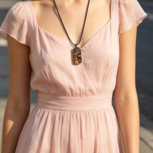 Puede incluir: Un collar con colgante de piedra rectangular estampada. La piedra presenta tonos marrones y beige. El collar est&aacute; suspendido de un cord&oacute;n negro. El colgante se lleva sobre un vestido rosa claro de manga corta.