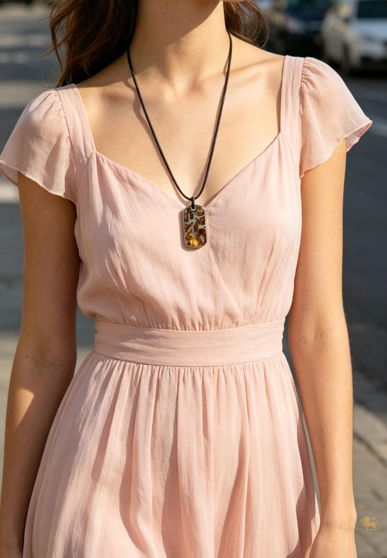 Puede incluir: Un collar con colgante de piedra rectangular estampada. La piedra presenta tonos marrones y beige. El collar est&aacute; suspendido de un cord&oacute;n negro. El colgante se lleva sobre un vestido rosa claro de manga corta.
