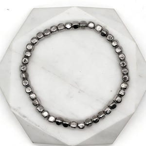 Puede incluir: Una pulsera de cuentas con pequeñas cuentas cuadradas de color gris oscuro. La pulsera se muestra sobre una superficie geométrica blanca. Las cuentas tienen un brillo metálico y la pulsera es circular.