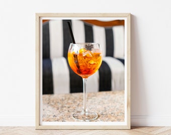 May include: A glass of orange and red cocktail with ice cubes and a black straw, sitting on a table with a granite-like surface. The background is blurred and shows a black and white striped couch.