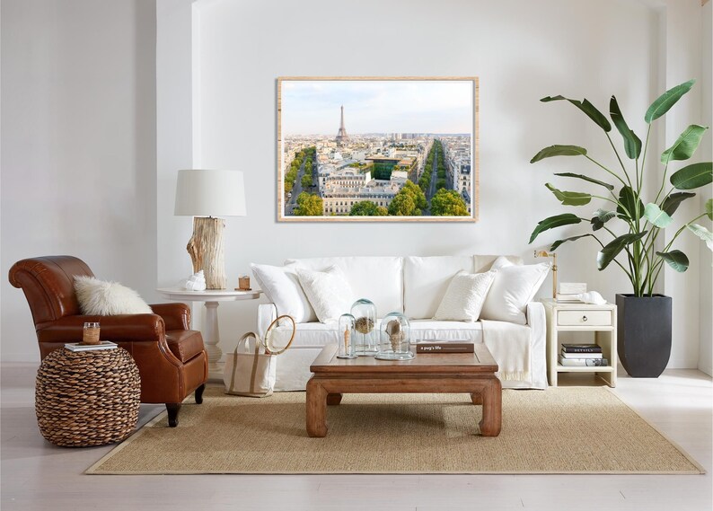 May include: A living room interior with a white sofa, a brown leather armchair, a wooden coffee table, and a framed print of the Eiffel Tower in Paris. The room is decorated with plants and other decorative items.