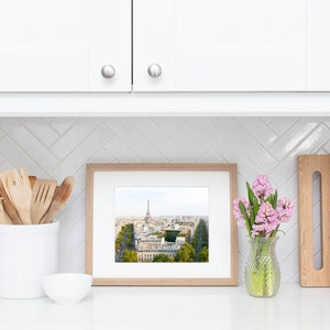 May include: A framed photograph of the Eiffel Tower in Paris, France, with a view of the city below. The photo is displayed on a white countertop with a white ceramic utensil holder, a white bowl, and a vase of pink flowers.