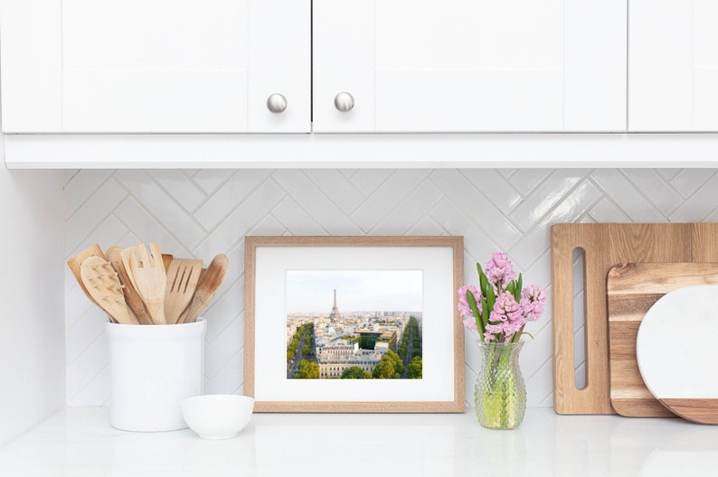May include: A framed photograph of the Eiffel Tower in Paris, France, with a view of the city below. The photo is displayed on a white countertop with a white ceramic utensil holder, a white bowl, and a vase of pink flowers.