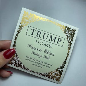 Pu&ograve; includere: Un'etichetta quadrata per il prodotto Trump Home Passion Citrus Soaking Salts. L'etichetta presenta un centro circolare verde pallido con testo nero, circondato da un bordo in foglia d'oro. Il testo recita: TRUMP HOME Passion Citrus Soaking Salts.