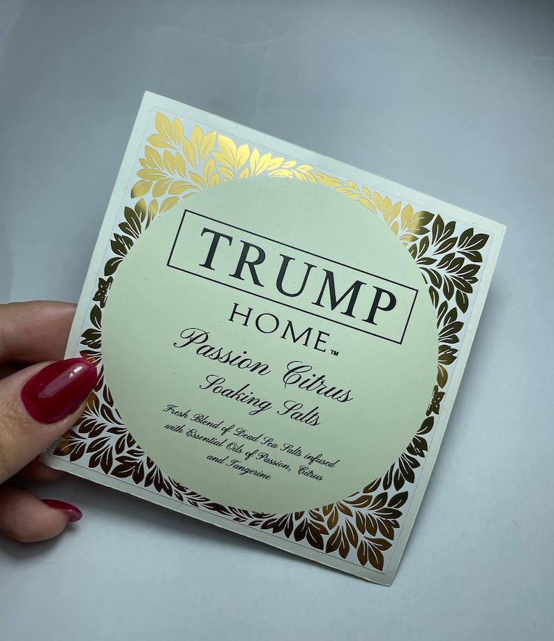 Pu&ograve; includere: Un'etichetta quadrata per il prodotto Trump Home Passion Citrus Soaking Salts. L'etichetta presenta un centro circolare verde pallido con testo nero, circondato da un bordo in foglia d'oro. Il testo recita: TRUMP HOME Passion Citrus Soaking Salts.