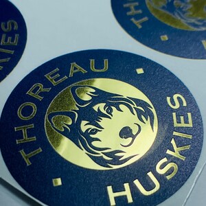 Pu&ograve; includere: Un adesivo circolare con un bordo blu scuro con il testo dorato THOREAU HUSKIES e un cerchio centrale dorato contenente un'illustrazione stilizzata di una testa di cane husky. Il design include piccoli rombi dorati che separano il testo.