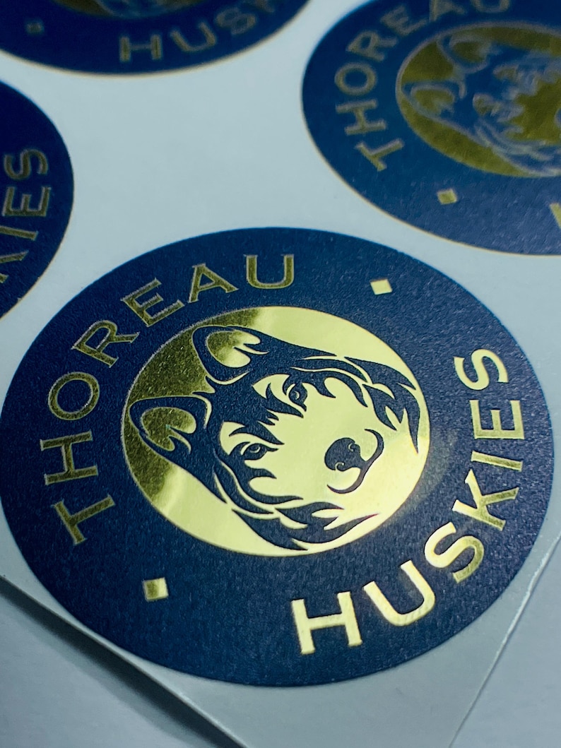 Pu&ograve; includere: Un adesivo circolare con un bordo blu scuro con il testo dorato THOREAU HUSKIES e un cerchio centrale dorato contenente un'illustrazione stilizzata di una testa di cane husky. Il design include piccoli rombi dorati che separano il testo.