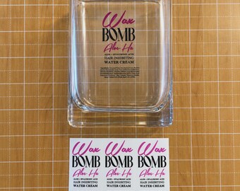 Può includere: Un contenitore rettangolare in vetro trasparente con il testo stampato Wax BOMB Aloe Ha HAIR INHIBITING WATER CREAM, accompagnato da tre etichette adesive rettangolari identiche che riportano lo stesso nome del prodotto e l'elenco degli ingredienti.