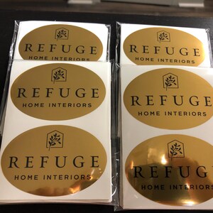 Pu&ograve; includere: Una collezione di adesivi ovali in lamina d'oro con un logo nero raffigurante il contorno di una casa con un ramo frondoso all'interno. Il testo REFUGE HOME INTERIORS &egrave; stampato in lettere maiuscole nere sotto il logo su ogni etichetta adesiva.