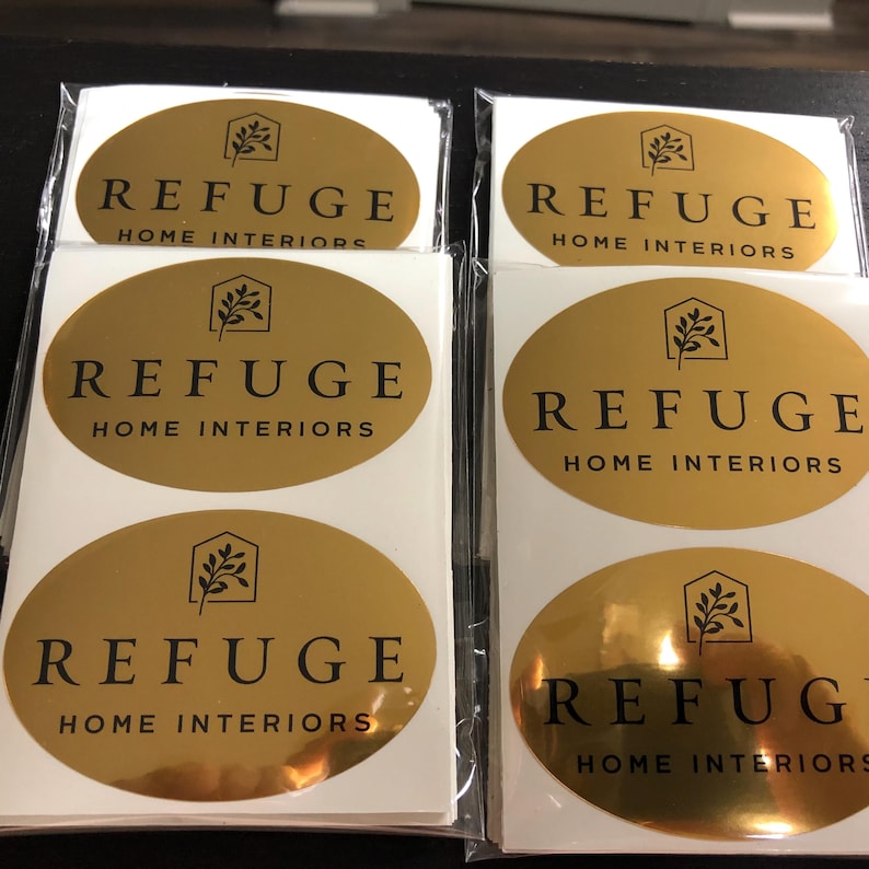 Pu&ograve; includere: Una collezione di adesivi ovali in lamina d'oro con un logo nero raffigurante il contorno di una casa con un ramo frondoso all'interno. Il testo REFUGE HOME INTERIORS &egrave; stampato in lettere maiuscole nere sotto il logo su ogni etichetta adesiva.