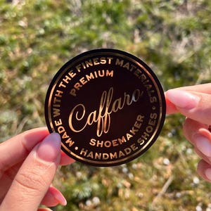 Pu&ograve; includere: Un adesivo circolare nero con scritte in lamina d'oro. Il testo riporta "Caffaro" in un carattere corsivo, circondato dalle parole "PREMIUM", "MADE WITH THE FINEST MATERIALS" e "SHOEMAKER HANDMADE SHOES" in un carattere sans-serif.