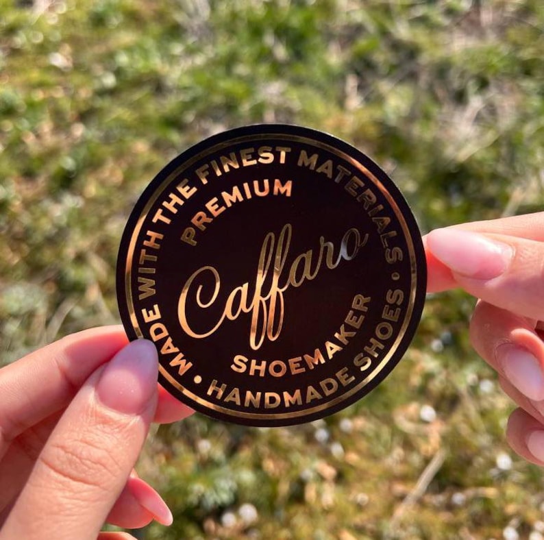 Pu&ograve; includere: Un adesivo circolare nero con scritte in lamina d'oro. Il testo riporta "Caffaro" in un carattere corsivo, circondato dalle parole "PREMIUM", "MADE WITH THE FINEST MATERIALS" e "SHOEMAKER HANDMADE SHOES" in un carattere sans-serif.