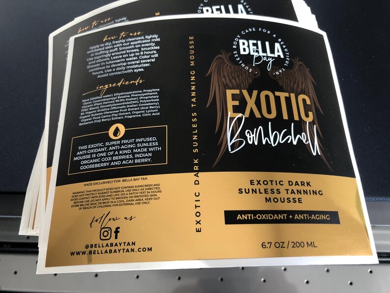 Pu&ograve; includere: Un'etichetta rettangolare per il prodotto Bella Bay Exotic Dark Sunless Tanning Mousse. Il design nero e oro presenta ali d'angelo, il nome del prodotto, ingredienti, istruzioni per l'uso e avvertenze. Include il testo 200 ML.