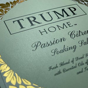 Pu&ograve; includere: Un'etichetta quadrata per prodotto con sfondo verde pallido e bordo in foglia d'oro. Il testo recita TRUMP HOME, Passion Citrus Soaking Salts, Fresh Blend of Dead Sea with Essential Oils of e Ta. Il design presenta un cerchio centrale.