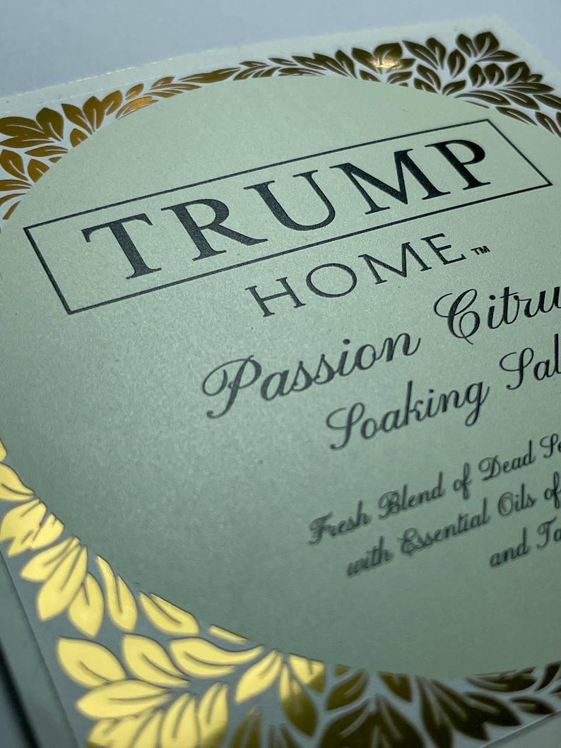 Pu&ograve; includere: Un'etichetta quadrata per prodotto con sfondo verde pallido e bordo in foglia d'oro. Il testo recita TRUMP HOME, Passion Citrus Soaking Salts, Fresh Blend of Dead Sea with Essential Oils of e Ta. Il design presenta un cerchio centrale.