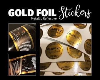 May include: Round gold foil stickers with a metallic reflective finish. The stickers feature the black text GLOW XO Sugar Scrub in a circular design. The image also shows black labels with gold text applied to a dark bottle, displaying Lorem Ipsum.