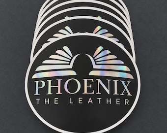 Può includere: Una pila di adesivi circolari con sfondo nero e bordo bianco. Il design include una grafica ad ali stilizzate olografiche sopra il testo PHOENIX THE LEATHER in un carattere sans-serif pulito, creando un effetto riflettente.