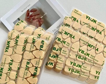 Può includere: Primo piano di diversi pacchetti di linguette adesive per unghie. Ogni linguetta è di colore beige chiaro con la scritta "YBJK Nail Adhesive" stampata in verde. Sullo sfondo c'è una rivista con le parole "World of Love".