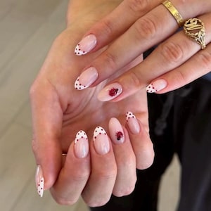 Pode incluir: Unhas em formato amêndoa com base rosa claro e pontas brancas salpicadas com pequenas bolinhas vermelhas. Algumas unhas apresentam um desenho de joaninha. Anéis dourados adornam os dedos.
