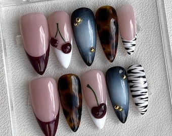 May include: A set of ten almond-shaped press-on nails in a clear plastic case. Designs include deep burgundy French tips, white tips with red cherry art, dark grey cat-eye polish with gold studs, brown tortoiseshell patterns, and black zebra stripes.