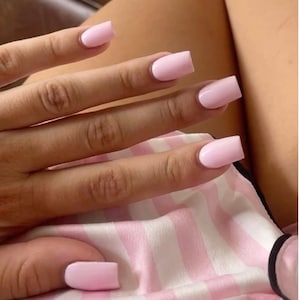 Può includere: Una mano con unghie acriliche rosa chiaro di forma quadrata. Le unghie sono di un colore pieno e opaco. Lo sfondo include un tessuto a righe rosa e bianche.