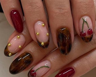 May include: Close-up of a hand with a manicure featuring various nail designs. Some nails are a deep red, others have a tortoiseshell pattern, and some are light pink with gold studs or cherry designs. The nails are oval-shaped.