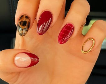 May include: A hand with five manicured nails. The nails feature a variety of designs, including a leopard print, solid burgundy, a red crocodile pattern, a gold ring, and a French tip with a burgundy crescent.