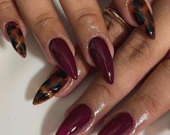 May include: Stiletto-shaped fingernails featuring a glossy finish. Some nails are painted in a solid deep burgundy color, while others display a brown and black tortoiseshell pattern. The nails are long, pointed, and have a smooth, reflective surface.