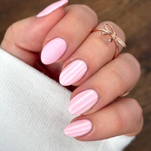 Könnte beinhalten: Nahaufnahme einer Hand mit ovalen, rosa lackierten Nägeln, einige mit weißen Streifen. Ein roségoldener Bogenring ziert einen Finger. Die Nägel sind hellrosa lackiert. Der Hintergrund ist eine Holzoberfläche.