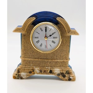 May include: A mantel clock with a textured tan sand finish and a deep blue arched top. The clock face is white with black Roman numerals, gold-tone trim, and three thin hands. The base is embedded with small pebbles and white seashells.
