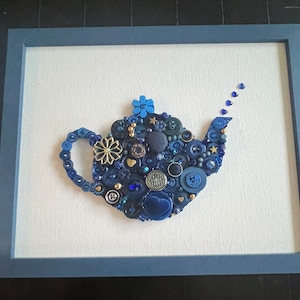 May include: A blue teapot made of buttons, beads, and other embellishments. The teapot is framed in a blue frame and is on a white background.