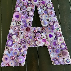 May include: A large letter A made of purple, pink, and white buttons. The letter is decorated with small flowers, hearts, and butterflies.