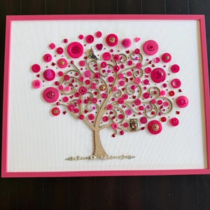 May include: A framed wall art piece featuring a tree silhouette with branches adorned with numerous pink buttons of varying sizes and shades. The tree includes a small metallic hummingbird and owl figure nestled among the pink button foliage.