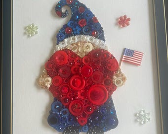 May include: Framed artwork of a gnome crafted from red, white, and blue buttons, holding a small American flag. The gnome's hat is blue, the body is red, and the beard is white. Decorative flower-shaped embellishments surround the gnome.