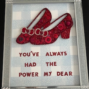May include: A framed artwork featuring a pair of red ruby slippers made from buttons. The background is a blue and white checkered pattern. The text "You've Always Had The Power My Dear" is written below the slippers.
