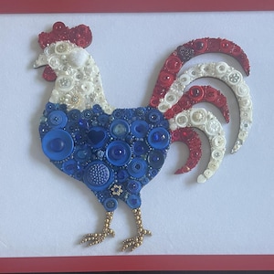 May include: A framed artwork featuring a rooster design crafted from various buttons. The rooster's body is blue, the head is white, and the comb and tail feathers are red. The rooster's feet are gold-colored. The artwork is set against a white background.