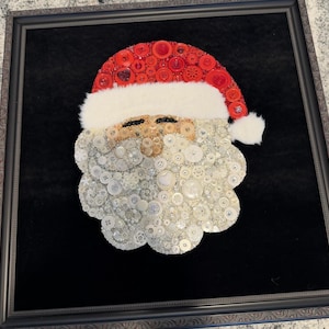 May include: Framed artwork featuring a Santa Claus face crafted from various buttons. The Santa hat is made of red buttons, the beard is white, and the face is tan. The artwork is set against a black background and framed in a decorative black frame.