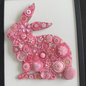 May include: A framed artwork featuring a pink bunny silhouette crafted from various pink buttons, beads, and embellishments. The buttons vary in size, shape, and shade, creating a textured and detailed design. The artwork is set against a white background within a black frame.
