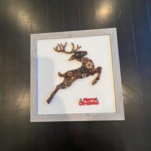 May include: A framed artwork featuring a reindeer made from buttons in shades of brown, gold, and red. The reindeer is jumping and has a red button for a nose. The artwork has a white background and the words "Merry Christmas" are written in red below the reindeer.