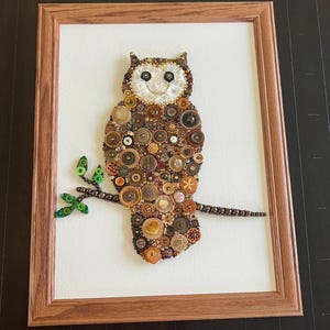 May include: A framed artwork featuring an owl made from buttons in shades of brown, beige, and gold. The owl is perched on a branch with green leaves.