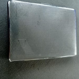 May include: A rectangular, silver-colored cigarette case with a textured, wave-like pattern. A diamond-shaped design is etched on the front. The case has a shiny, metallic finish and a hinged lid.