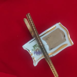 以下が含まれることがあります： 金色の縁取りと金色の金属製のポールが付いた、小さな装飾用の磁器製の旗ピン。旗は白で、金色の長方形と紫色の花の模様があり、赤い背景に配置されています。