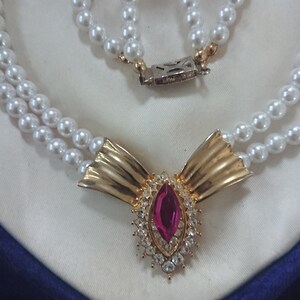 May include: A double-strand pearl necklace with a gold-toned bow-shaped pendant. The pendant features a large, oval, pink gemstone surrounded by clear crystals. The necklace is displayed on a white satin surface.