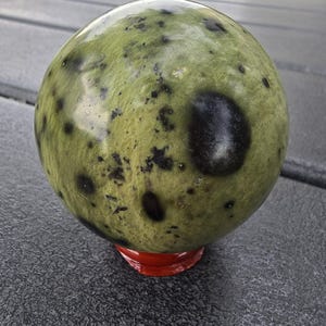 May include: A large, spherical decorative stone with a mottled green and black pattern. The stone sits on a small, round, red base, and is displayed on a gray surface. The stone's surface has a polished, glossy appearance.